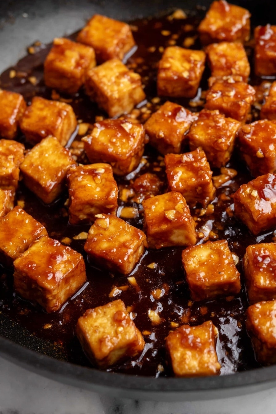 orange tofu - The image shows many small, golden brown tofu cubes in a black pan, covered with a thick, shiny, dark brown sauce with small bits of garlic or seasoning scattered in it. The tofu cubes have a slightly crispy texture on the outside and are evenly coated with the sauce, which pools a little at the bottom of the pan. The background is a white marbled surface. photo taken with an iphone --ar 2:3 --v 7