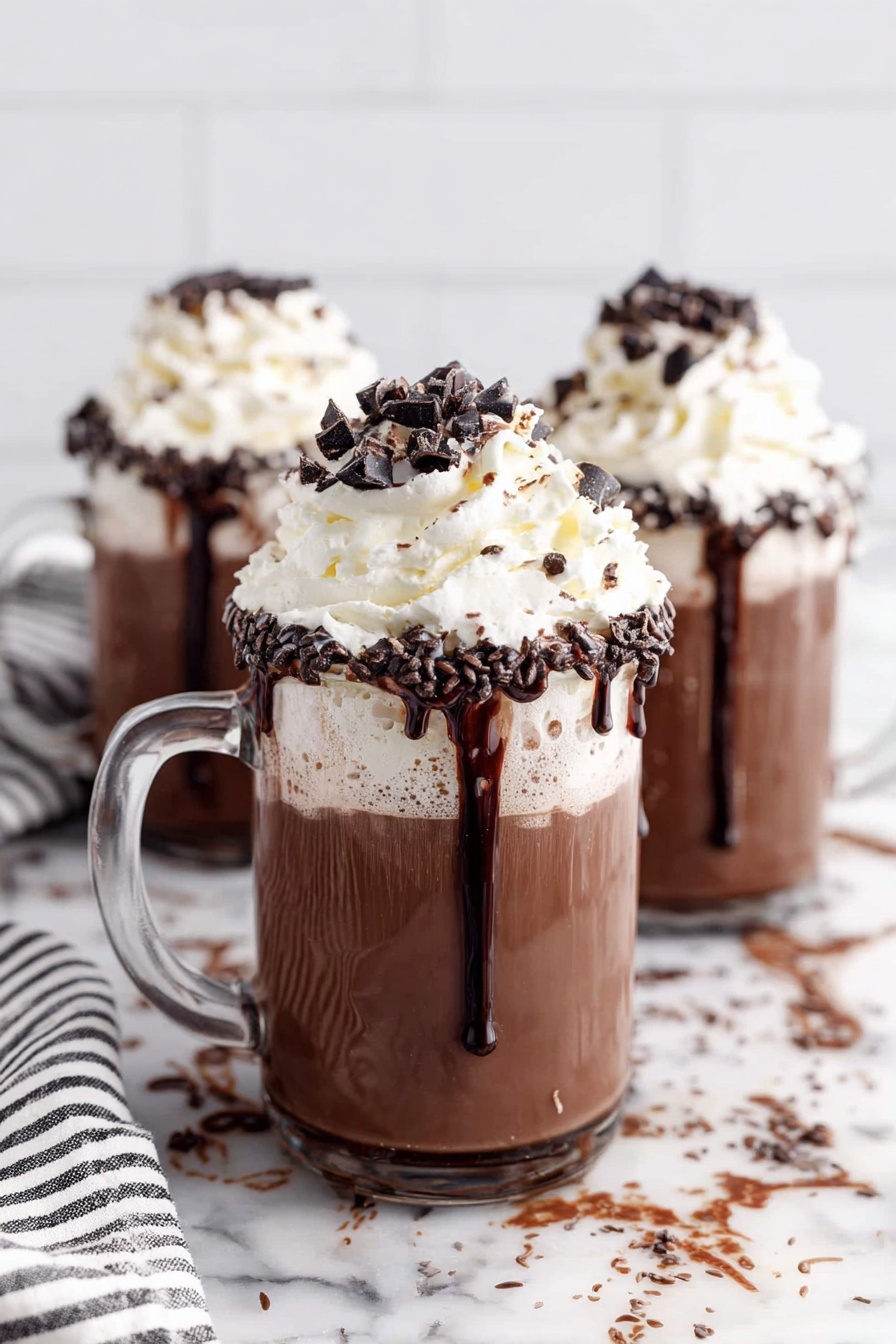 The image shows a glass jar mug filled with three layers: a light brown creamy base layer at the bottom, topped with a lighter whipped cream layer in the middle, and a large puff of white whipped cream on top sprinkled with dark chocolate chips and small chocolate sprinkles. The rim of the jar is covered with a thick layer of dark chocolate sprinkles. The mug has a clear handle and is placed on a white marbled surface. In the blurry background, there are two more similar glass jar mugs with the same drink. Photo taken with an iphone --ar 2:3 --v 7