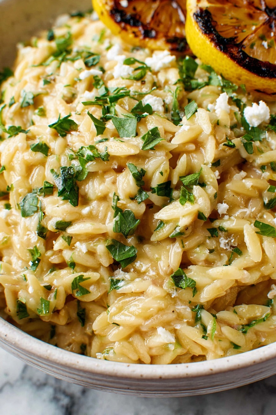 The image shows a close-up view of creamy orzo pasta in a white bowl, with a rich texture and light golden color mixed with small green herb pieces scattered on top. There are some small crumbles of white cheese and finely chopped fresh herbs like parsley or basil on the surface, adding green and white spots to the creamy yellow base. In the background, a charred grilled lemon half is visible, with caramelized dark lines contrasting with the bright yellow of the lemon. The bowl rests on a white marbled surface. photo taken with an iphone --ar 2:3 --v 7