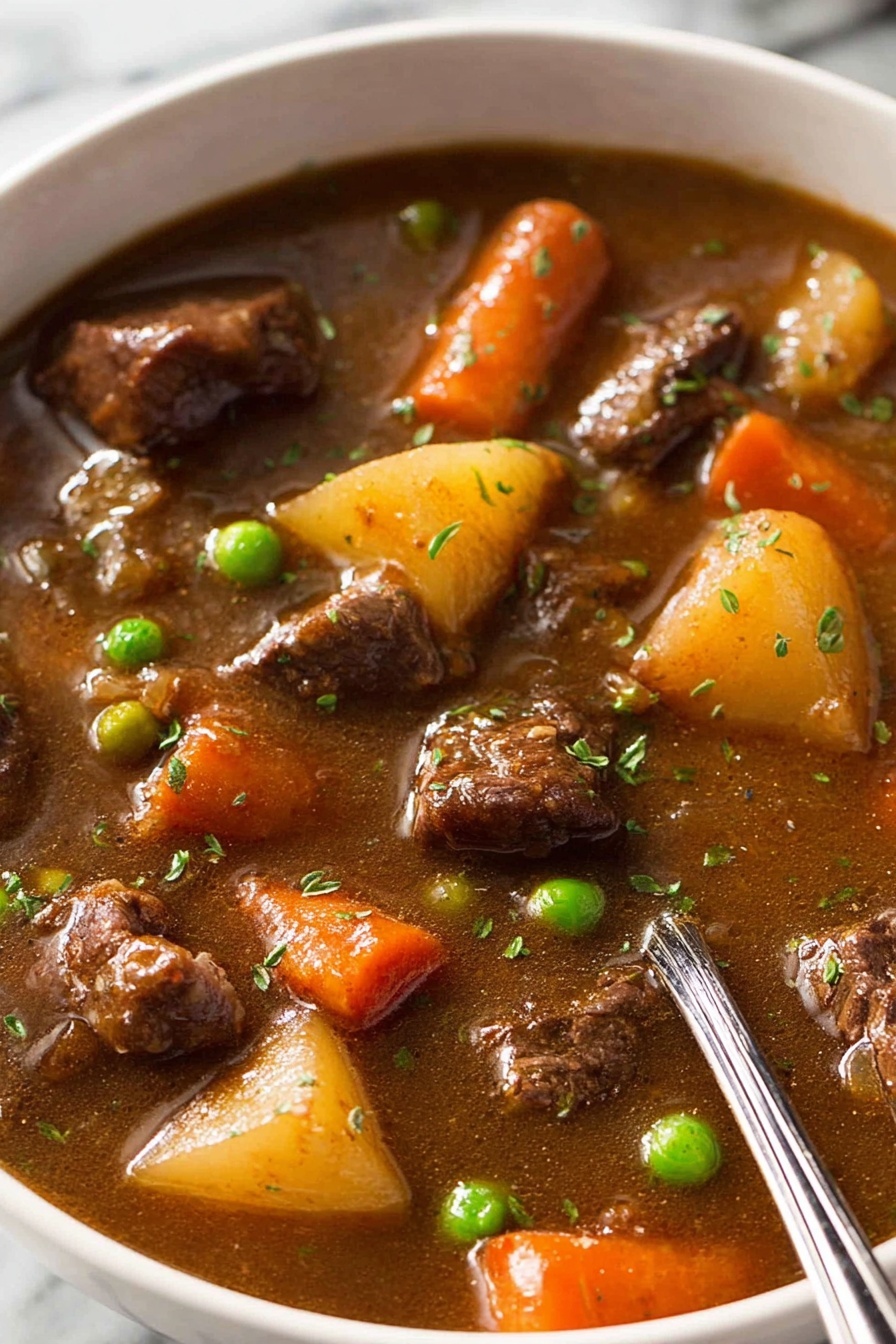 vegetable beef soup - A close-up view of a thick stew filled with several layers of chunky ingredients floating in a rich brown broth. The stew has medium-sized pieces of tender dark brown meat scattered throughout, bright orange carrot pieces, green peas, chunks of light yellow potatoes, and bits of celery, all mixed with small green herb sprinkles on top. The liquid looks smooth and slightly shiny, covering everything evenly. A silver spoon rests inside the bowl from the right edge. The bowl is white, sitting on a white marbled surface. Photo taken with an iphone --ar 2:3 --v 7