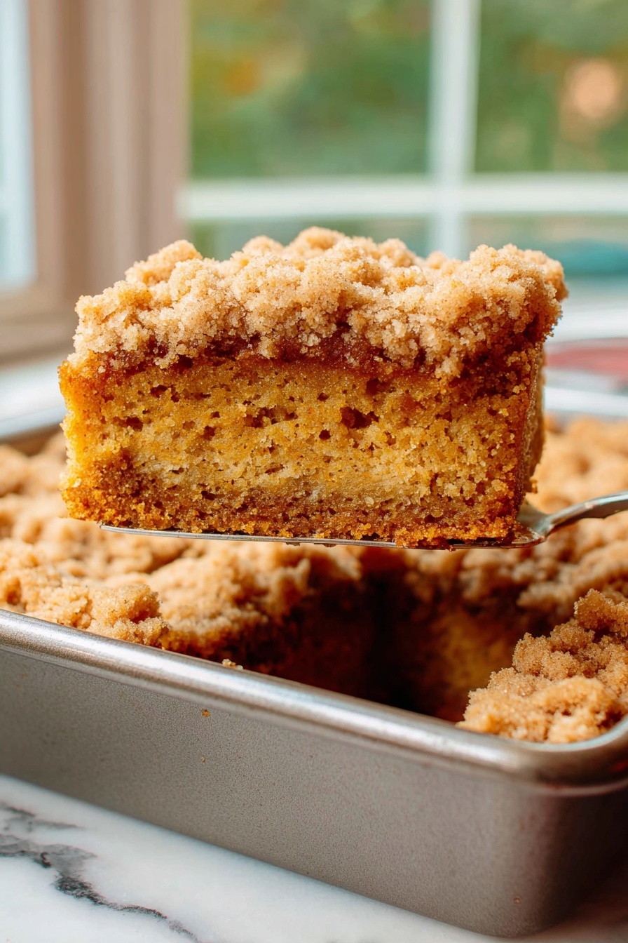 A thick square piece of crumb cake is lifted above a metal baking pan filled with more crumb cake. The cake has two main layers: a dense, moist, orange-brown bottom layer with a soft texture full of small holes, and a top layer made of light brown, crumbly streusel with a rough texture. The crumb layer sits evenly on the cake but is uneven on top, giving a rustic look. The background shows a blurred window and green outside. The photo is taken on a white marbled surface. Photo taken with an iphone --ar 2:3 --v 7
