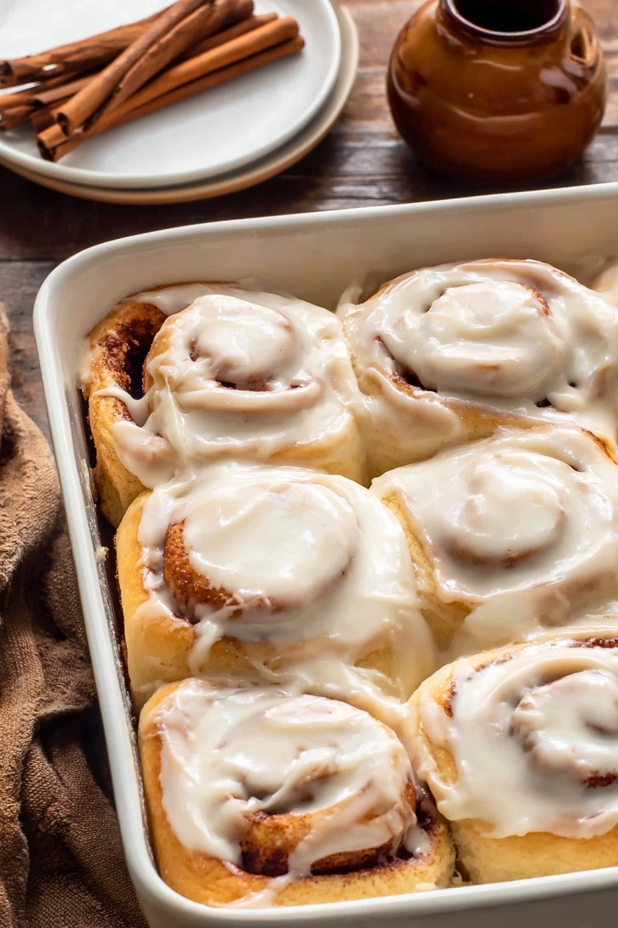 A close-up of a white rectangular baking dish filled with four cinnamon rolls arranged in two rows, each roll showing thick, creamy white icing spread unevenly on top, covering the soft tan dough with visible darker cinnamon swirl layers inside. The dish is placed on a dark wood surface with a round white plate holding cinnamon sticks and a small brown ceramic jar nearby. Photo taken with an iphone --ar 2:3 --v 7