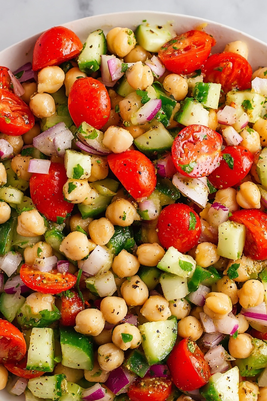 The image shows a close-up of a chickpea salad with multiple layers of ingredients mixed together. The dish has a base layer of light beige chickpeas that are round and smooth. Among the chickpeas are bright red cherry tomato halves with a shiny, juicy texture placed all around. There are chopped cucumber pieces in pale green with darker green skin edges scattered evenly. Small chunks of light purple-red onion and dark green bell pepper cubes are mixed in, adding texture and color contrast. The salad is garnished with small flecks of chopped green herbs and some black pepper seasoning visible throughout. The whole salad is sitting on a white marbled surface photo taken with an iphone --ar 2:3 --v 7