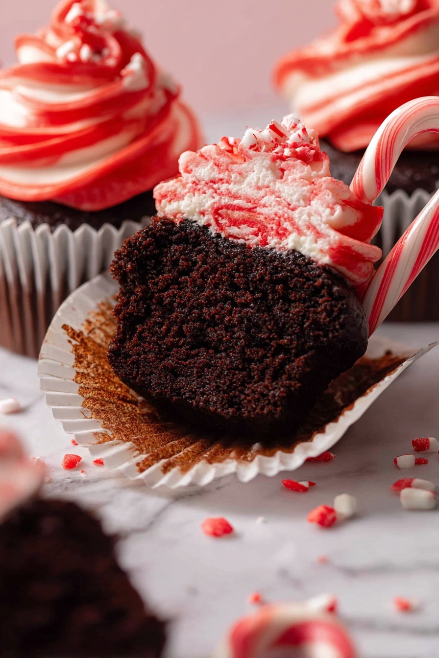 A close-up view of dark chocolate cupcakes with two layers visible: the moist, dark brown cake base and a thick swirl of red and white striped frosting on top. One cupcake is cut in half, showing its soft interior, with one half still in the white cupcake liner and the other lying on the white marbled surface. Small red and white candy pieces are scattered around the cupcakes, and a small red-and-white striped candy cane sticks out from the frosting on the cut cupcake. The background is a soft white marbled texture. photo taken with an iphone --ar 2:3 --v 7
