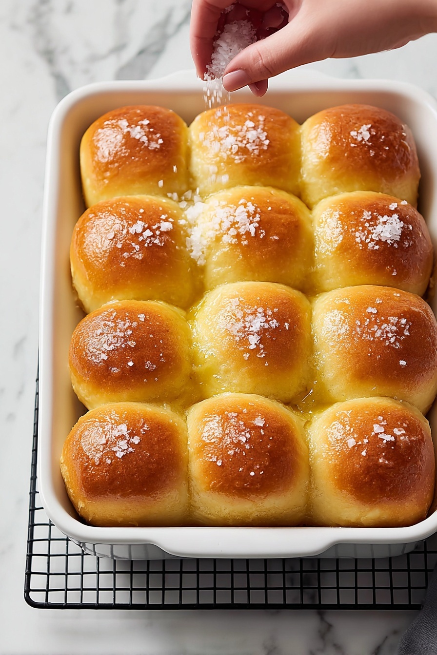 A single soft bread roll sits on a small white plate, showing a torn side that reveals its fluffy, light beige interior texture. The top of the roll is golden brown with some coarse sea salt sprinkled on it, giving a slight shine. Behind the plate is a white baking dish filled with more bread rolls, each with a similar golden-brown top. To the right of the plate, a small white bowl holds two pale yellow butter slices. The whole scene is set on a white marbled surface with bright, natural lighting. Photo taken with an iphone --ar 2:3 --v 7