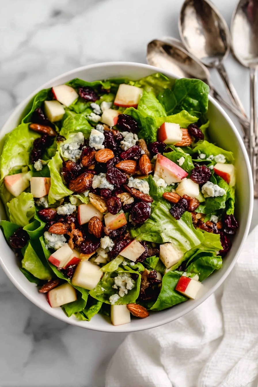 The dish shows a white bowl filled with fresh green lettuce leaves as the base layer, topped with small red and white apple cubes scattered all over. On top of that is a layer of roasted brown almonds and dark red dried cranberries evenly spread. The final layer includes small chunks of blue cheese dotted across, creating a mix of green, red, brown, and creamy white colors with different textures. The bowl sits on a white marbled surface with a white cloth nearby and two silver spoons to the top right. photo taken with an iphone --ar 2:3 --v 7
