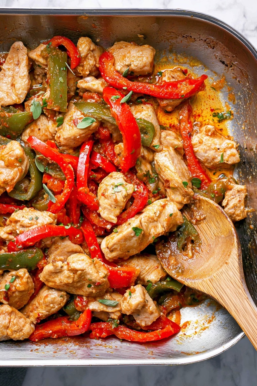 A close-up view of a metal pan filled with cooked chicken pieces and sliced bell peppers. The chicken pieces are light brown with a slight sear and mixed with red and green bell pepper strips that are soft and shiny from cooking. Some small green herb leaves are scattered on top, and a wooden spoon with a long handle rests inside the pan, partially mixing the ingredients. The pan surface has some sauce residue with a creamy orange tint, showing the cooked bits stuck to the bottom. The background features a white marbled texture. Photo taken with an iphone --ar 2:3 --v 7