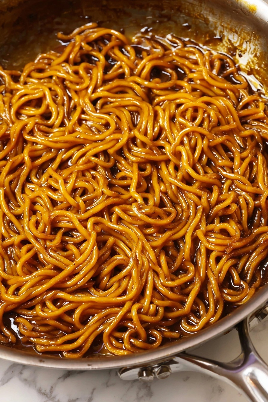 A close-up view of cooked noodles inside a metal frying pan. The noodles are coated in a shiny, dark brown sauce that makes them look smooth and slippery. The pan's inner surface is dark and shows some sauce residue. The metal handle of the pan is partly visible with a simple screw holding it in place. The background shows a white marbled texture beneath the pan. photo taken with an iphone --ar 2:3 --v 7