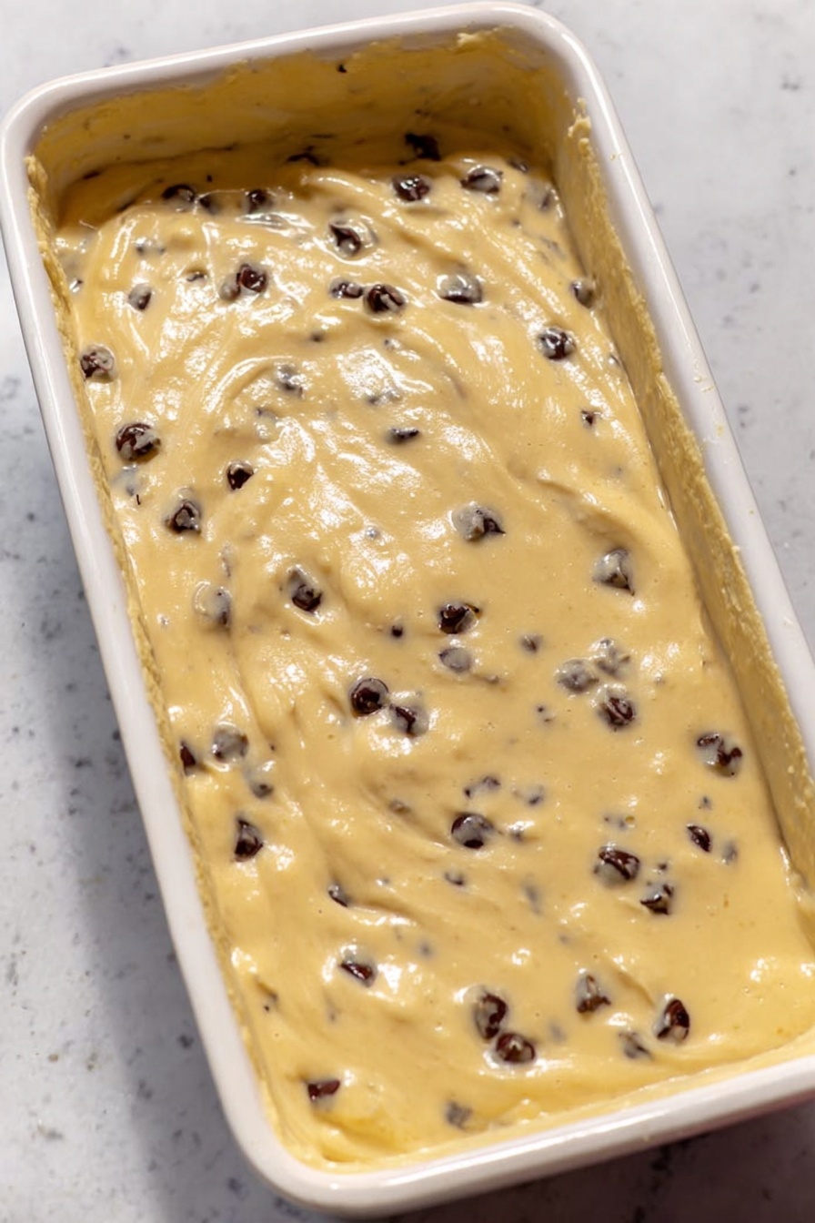 A slice of brown banana bread with dark chocolate chips mixed inside is placed on a white plate with a fork next to it. The slice has a thick layer of smooth, orange-tan frosting on top, sprinkled with small dark chocolate chips and small bits of candied orange peel. Behind the plate, the rest of the loaf sits on a metal tray, showing the same texture inside and covered with the same frosting and toppings. The background features a white towel with blue stripes, a yellow banana, and small white bowls with chocolate chips and candied orange pieces on a white marbled surface. photo taken with an iphone --ar 2:3 --v 7