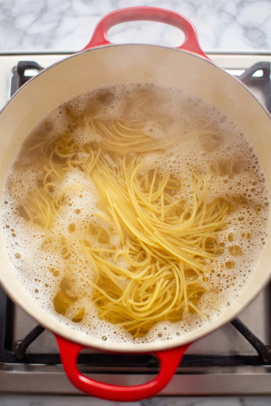 A bowl filled with three layers: the bottom layer is a black bowl with a smooth mat finish; the middle layer includes light brown noodles twisted in soft loops and mixed with small golden brown nuts and several dark red dried whole chili peppers placed around the edges; the top layer consists of shiny golden brown bite-sized chunks of coated chicken scattered among the noodles, bright green chopped scallions dotted on top, and a fork with a vintage silver handle resting inside the bowl partly under the noodles. The bowl is placed on a white marbled surface with a small wooden bowl of green scallions in the top right corner and a red cloth napkin peeking from the bottom left corner. photo taken with an iphone --ar 2:3 --v 7