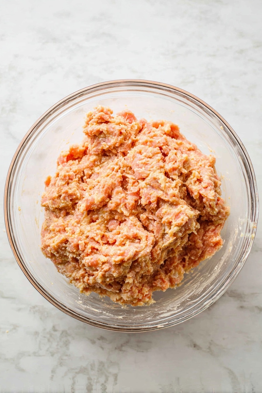 A white bowl filled with round meatballs covered in a creamy light beige sauce with visible black pepper and green herb bits. The meatballs are mixed with small pieces of red sun-dried tomatoes and fresh green basil leaves scattered on top. A golden fork is lifting one meatball, showing the creamy texture and herbs inside. The background surface is white marble, and a corner of a rust red cloth is visible. Photo taken with an iphone --ar 2:3 --v 7